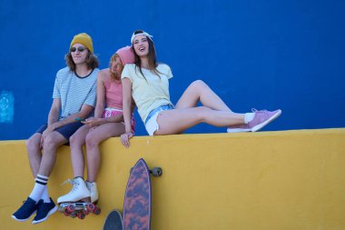 Canlı sarı bir duvarda oturan üç arkadaş çarpıcı bir mavi arka plana karşı, boş zamanların tadını çıkarıyor ve birlikte neşeli anları paylaşıyorlar.