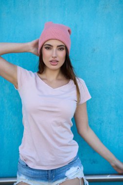 Pembe bereli, tişörtlü ve kot şortlu şık genç bir kadın, canlı mavi bir duvara karşı poz veriyor.