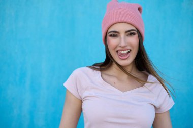 Pembe bereli ve tişörtlü genç bir kadın, parlak mavi bir arka plana karşı dilini uzatıyor.