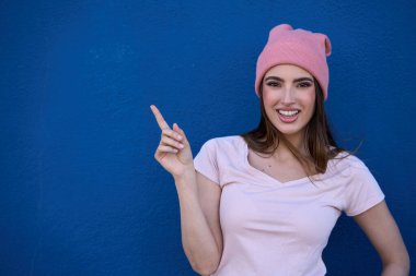 Pembe yün şapkalı, gülümseyen genç bir kadın, parlak mavi bir arka plana parmağını göstererek, pozitif ve neşe saçıyor.