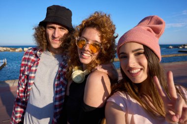 Üç mutlu arkadaş deniz kenarında selfie çekiyor, gülümsüyor ve barış işareti yapıyor, açık havada güneşli bir günün tadını çıkarıyorlar.