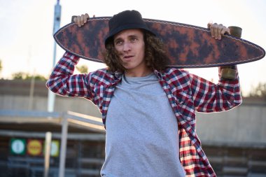 Kıvırcık saçlı kaykaycı omuzlarında uzun bir kaykayla gün batımında canlı bir kaykay parkına bakıyor.