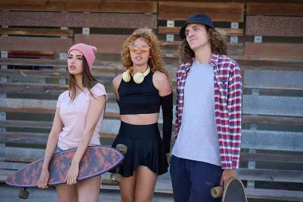 Metal örgülü ahşap bir duvara poz veren, kentsel gençlik kültürü ve kaykay yaşam tarzını gözler önüne seren üç havalı genç.