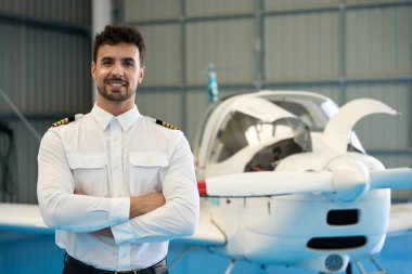 Kollarını kavuşturup gülümseyen kendinden emin bir pilotun portresi bir uçak hangarında küçük bir özel uçağın önünde gururla duruyor.
