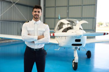 Kollarını kavuşturup gülümseyen kendinden emin pilot hangardaki küçük bir uçağın önünde gururla poz veriyor, profesyonelliği ve başarıyı simgeliyor.