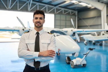 Kendine güvenen bir pilotun portresi havacılık hangarında uçakların önünde çapraz kollarla duruyor, havacılık endüstrisinde profesyonellik ve uzmanlık gösteriyor.