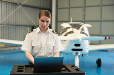 Dizüstü bilgisayar kullanan kendine güvenen kadın pilot, uçuş öncesi kontrolleri ya da bakım görevlerini hangarda, arka planda küçük özel uçakla yapıyor.