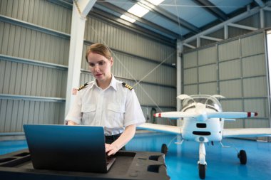 Dizüstü bilgisayarda çalışan kendinden emin bir kadın pilot, yakınlarda özel bir jetle bir hangarın içinde titizlikle uçuş planı hazırlıyor.