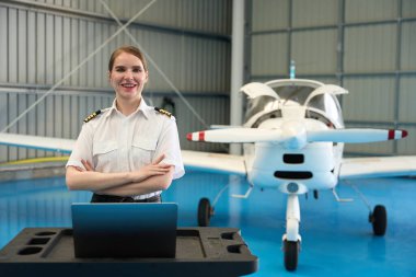 Genç kadın uçak pilotu kollarını kavuşturup gülümsüyor, hangarda küçük özel bir uçağın önünde duruyor.
