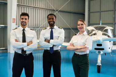 Hangarda küçük bir uçağın önünde kollarını kavuşturmuş gülümseyen öğrenci pilotların portresi.