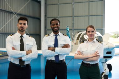 Üç gülümseyen pilot, bir uçak hangarında küçük bir uçağın önünde kollarını kavuşturarak duruyorlar.