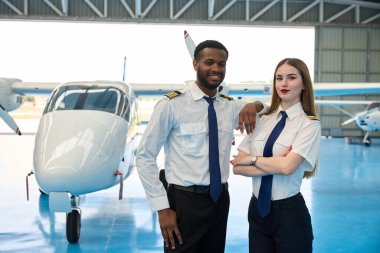 İki gülümseyen pilot, bir uçak hangarında küçük bir yolcu uçağının önünde kollarını kavuşturarak poz veriyorlar.
