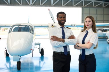 Kendine güvenen iki pilot, bir uçak hangarında küçük bir uçağın önünde kollarını kavuşturarak poz veriyor.