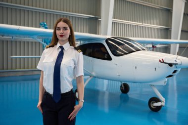 Bir havacılık hangarında küçük bir uçağın önünde gururla duran genç bayan pilot, havacılık endüstrisindeki kariyer isteklerini temsil ediyor.