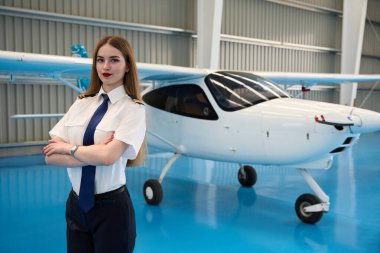 Genç kadın pilot havacılık hangarında küçük bir uçağın önünde çapraz kollarla duruyor. Geleceğin havacılığını temsil ediyor.