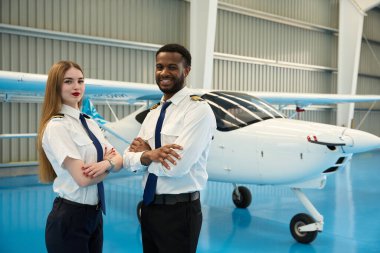 Havacılık okulunun hangarında iki gülümseyen pilot kollarını kavuşturup küçük bir uçağın önünde poz veriyor.