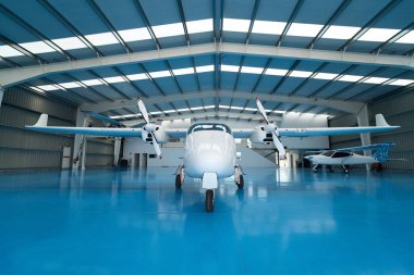 Mavi zemin ve diğer uçaklarla birlikte havacılık okulundaki hangarın içine park edilmiş beyaz çift motorlu uçak.