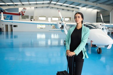 İş kadını, içi gösterişli küçük uçaklarla dolu bir uçak hangarına girerken kendinden emin bir şekilde bir tramvay bavulu itiyor.