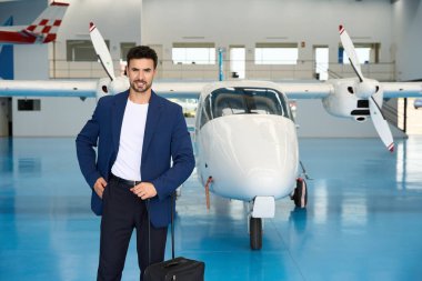 Tramvay çantası tutan bir iş adamı, lüks bir seyahat somutlaştıran lüks bir hangarın içinde, özel bir uçağın önünde güven içinde poz veriyor.
