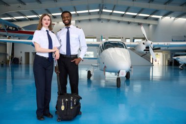 Arka planda hafif bir uçakla hangarda poz veren gülümseyen iki genç pilot öğrenci eğitim uçuşuna hazır.