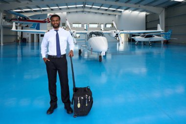 Uçak hangarında duran kendinden emin bir pilot. Arka planda tramvay valizi ve uçakları var. Havacılık uzmanı seyahat etmeye hazır.