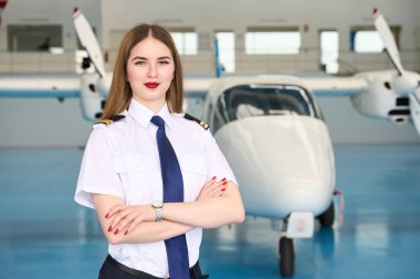 Hangarda küçük bir yolcu uçağının önünde çapraz kollu duran genç kadın pilot.