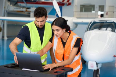 Güvenlik yelekleri giyen iki uçak tamircisi bir uçak hangarında uçak bakımı için dizüstü bilgisayar kullanıyor.