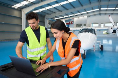 Güvenlik yelekleri giyen iki uçak tamircisi, arka planda küçük bir uçak bulunan bir hangarda dizüstü bilgisayar kullanıyorlar.