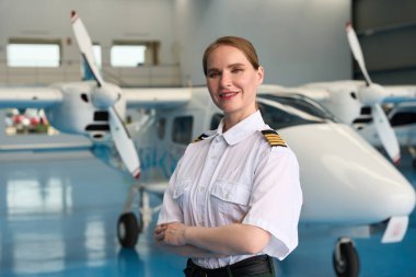 Bir uçak hangarında iki küçük uçağın önünde kollarını kavuşturmuş gülümseyen bir kadın pilotun portresi.