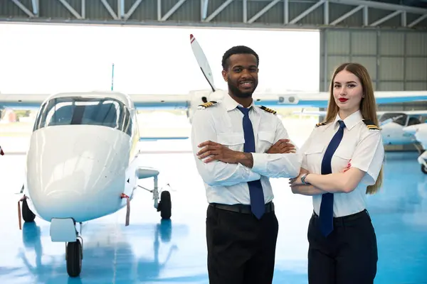 Kendine güvenen iki pilot, bir uçak hangarında küçük bir uçağın önünde kollarını kavuşturarak poz veriyor.