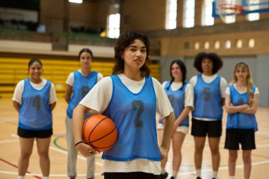 Genç bayan basketbolcu, arka planda takımıyla birlikte sahada top tutuyor. Maç için hazır.