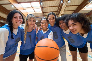 Mutlu basketbol oyuncuları bir top tutuyor, gülümsüyor ve canlı bir spor salonunda birlikte poz veriyorlar, takım çalışmasını ve arkadaşlığını kutluyorlar.