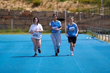 Üç genç kadın mavi bir pistte koşuyor, azim ve atletizm sergiliyorlar.