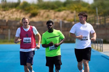Bir spor müsabakası sırasında mavi pistte koşan üç erkek sporcu kararlılık ve atletizm gösteriyor.