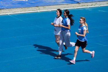 Üç genç kadın mavi pistte bir yarış düzenliyor, bir spor müsabakasında atletizm ve kararlılıklarını sergiliyorlar.