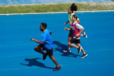 Bir atletizm müsabakası sırasında mavi pistte koşan üç erkek sporcu hız ve kararlılıklarını sergiliyorlar.