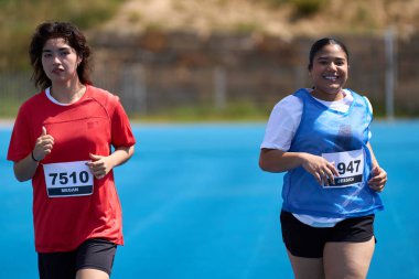 İki bayan sporcu mavi bir pistte koşu yaparken azim ve sportmenlik gösteriyorlar.