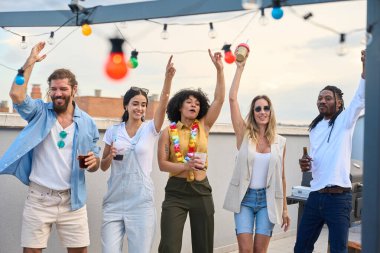 Mutlu çoklu etnik arkadaşlar gülüyor, dans ediyor ve canlı bir çatı partisinde içkilerini yudumluyor, yaz akşamlarını neşeyle kutluyorlar.