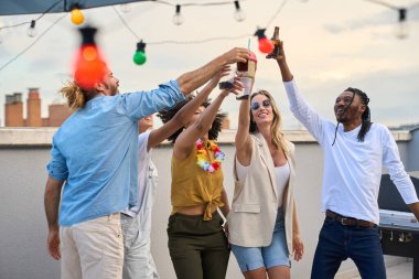 Neşeli çoklu etnik arkadaşlar kadeh kaldırıyor, kadeh kaldırıyor, çatı katındaki bir partide kadeh kaldırıyor, içkilerin tadını çıkarıyor ve arkadaşlıklarını kutluyorlar.