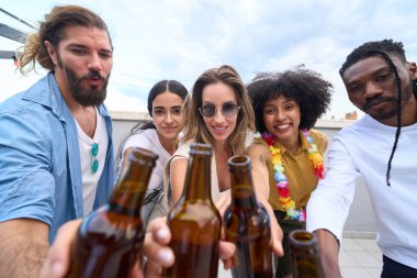 Neşeli çoklu etnik arkadaşlar çatı partisinde eğleniyor, kutlama için bira şişelerini kaldırıyorlar.