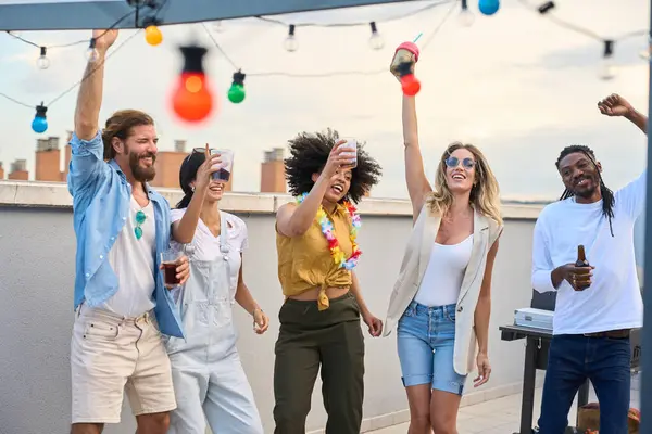 Neşeli çoklu etnik arkadaşlar bir çatı partisinde dans ediyor, içiyor ve kadehlerini kaldırıyorlar.
