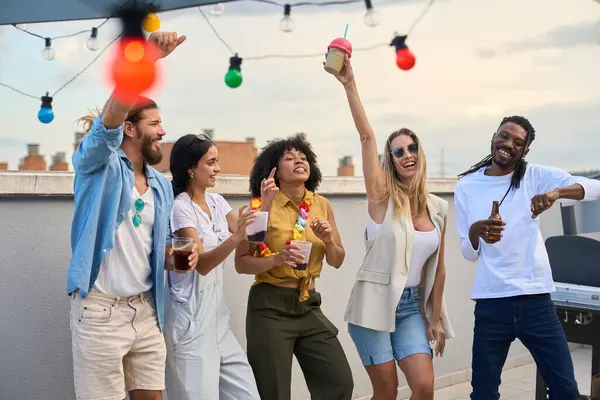 Neşeli çoklu etnik arkadaşlar dans ediyor ve içkilerini yudumluyorlar. Güneş batarken çatıdaki partinin coşkulu atmosferinin tadını çıkarıyorlar.