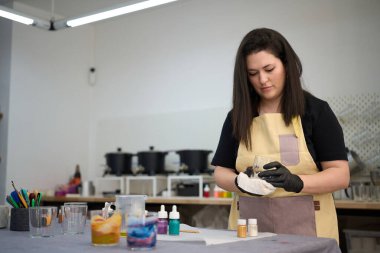 Önlük ve eldiven giyen bir kadın renkli bir stüdyoda mum yapmak için cam hazırlıyor, balmumu ve aletler kullanıyor.