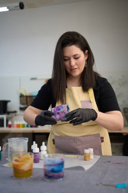 Önlük ve siyah eldiven giyen kadın sanatçı renkli zanaat atölyesindeki el yapımı mumu dikkatle inceliyor.