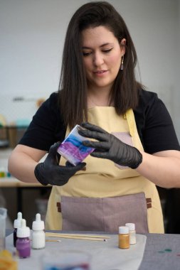 Eldiven takan usta kadın atölyesinde bitirilmiş bir reçine mumunu dikkatlice inceliyor, renkli pigmentler ve aletlerle çevrili.