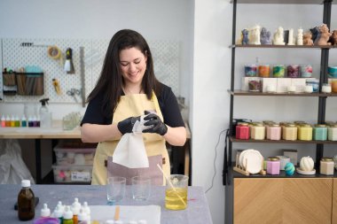 Cam kavanozu temizleyen gülümseyen usta kadın renkli atölyesinde el yapımı mumlar yapmak için balmumu dökmeye hazırlanıyor.