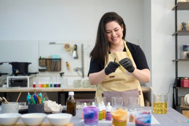 Mum yapımında çalışan bir kadın, renkli bir atölyede balmumu çarşaflar hazırlıyor. Etrafı alet ve malzemelerle çevrili.