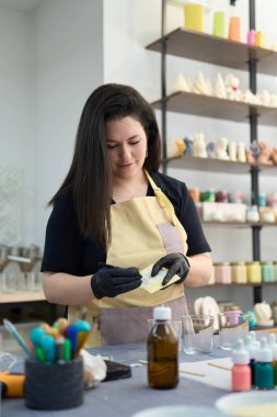 Odaklanmış usta fırça ve eldiven kullanarak balmumu hazırlıyor, renkli tamamlanmış ürünler ve aletlerle çevrili canlı atölyesinde eşsiz mumlar yaratıyor.