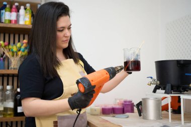 Sanatçı kadın ısı tabancası kullanarak renkli balmumu şişesini ısıtıyor ve parlak stüdyosundaki mum kalıbına dökmeye hazırlanıyor.