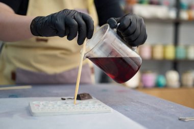 Yakın plan çekimde, eldivenli bir zanaatkar canlı stüdyoda el yapımı mumlar yaparak beher kabından kırmızı mum döker.